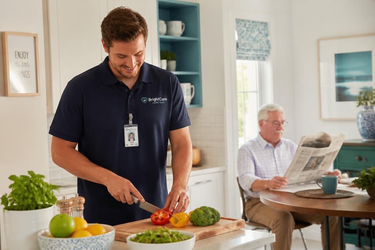 Caregiver preparing a healthy meal while an elderly gentleman reads a newspaper